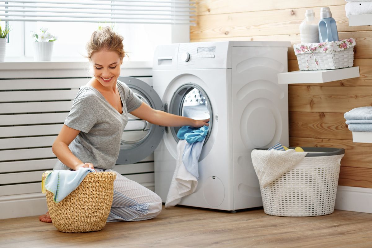 A person crouches beside a modern washing machine, loading it with colorful laundry while surrounded by cozy home decor.