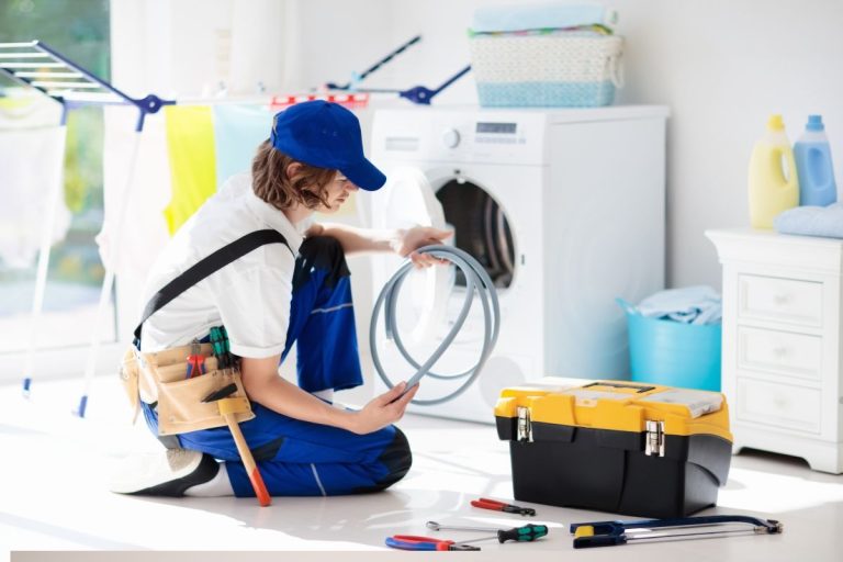 A technician kneels beside a washing machine, holding a hose, with a toolbox and various tools scattered on the floor. Bright laundry in the background.