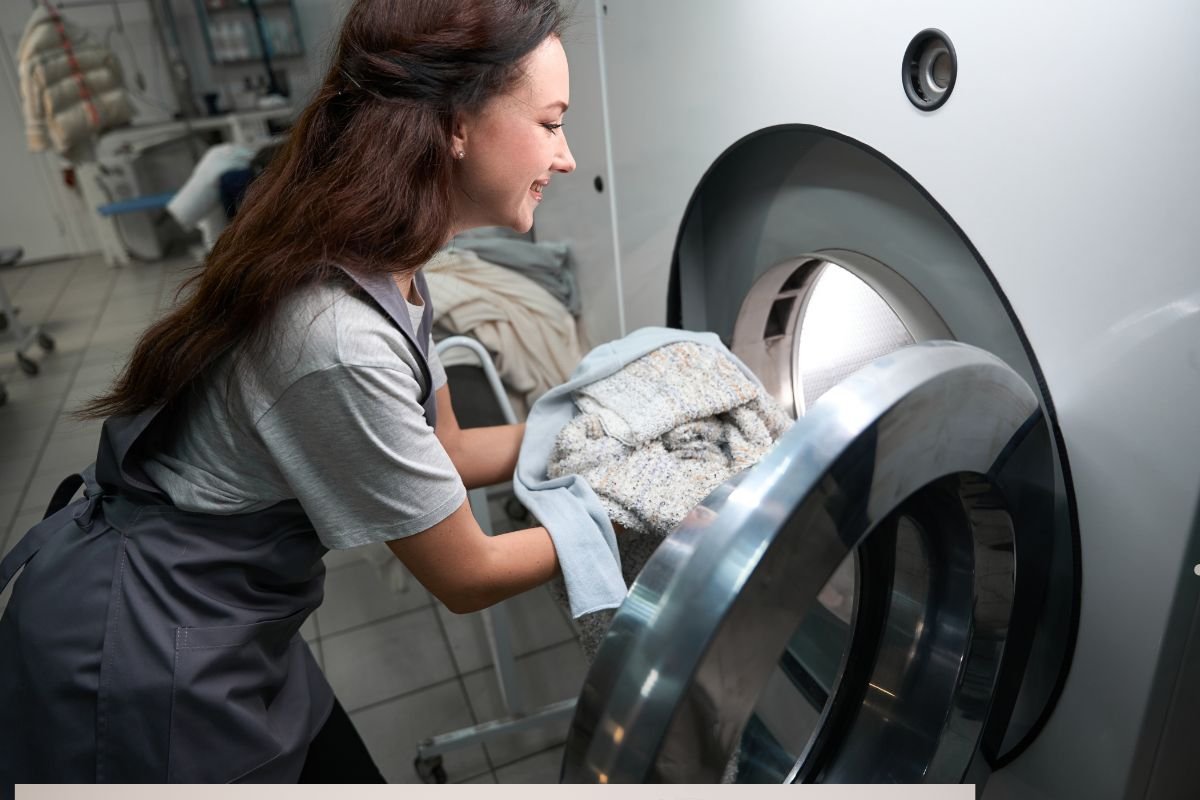 A person in a gray shirt is loading a large washing machine with folded towels and blankets in a laundry facility.